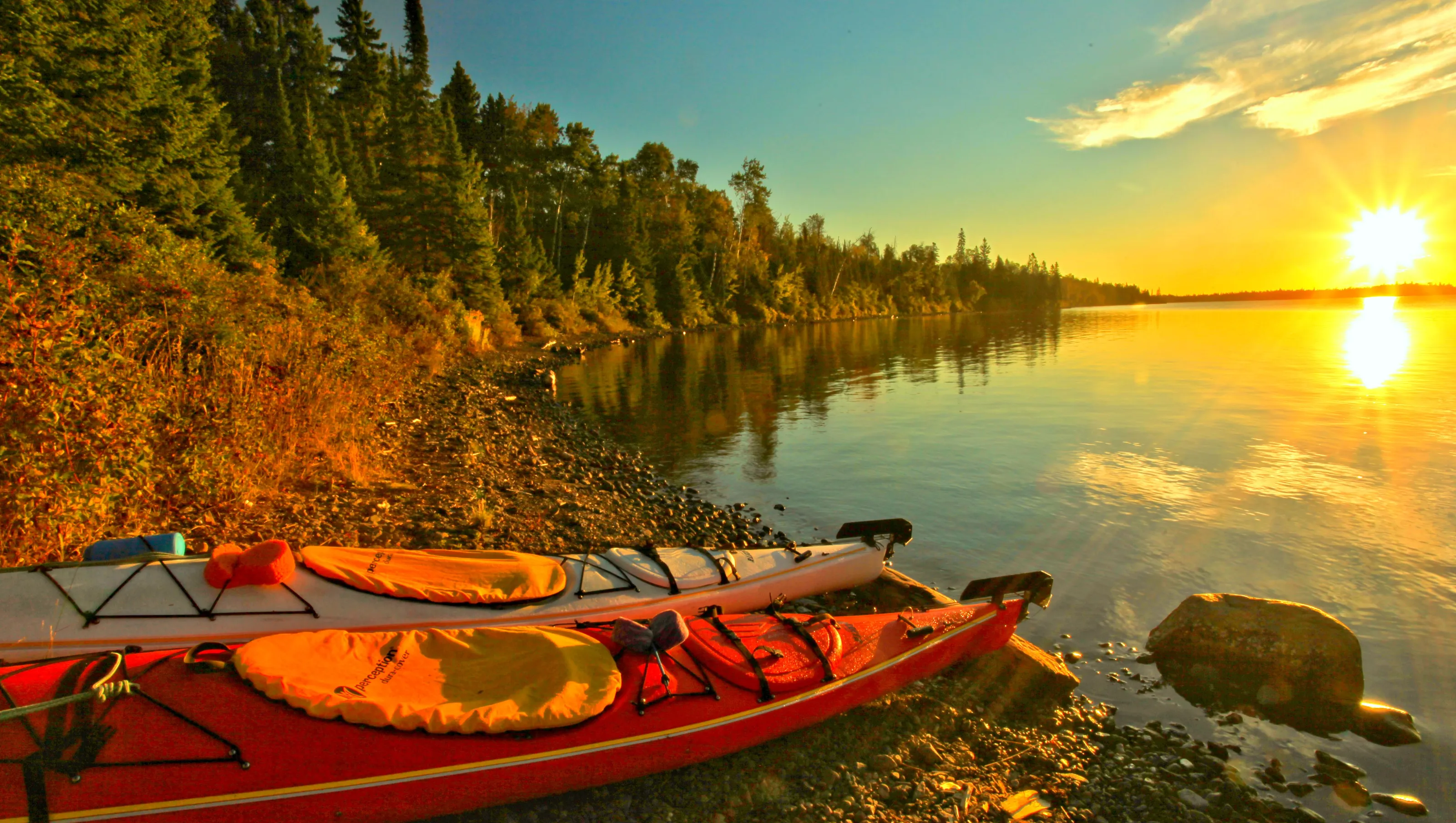 Two kayaks on the shore line with a sunset in the distance and a tree line going past the kayaks