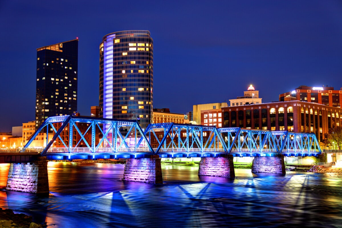 Bridge lit up in blue and purples at night time with large skyscrapers in the distance. 